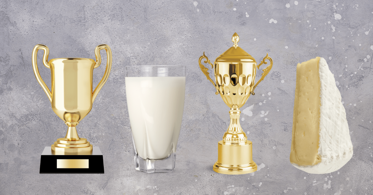 Two gold trophies lined up with a glass of milk and a triangle of brie cheese.